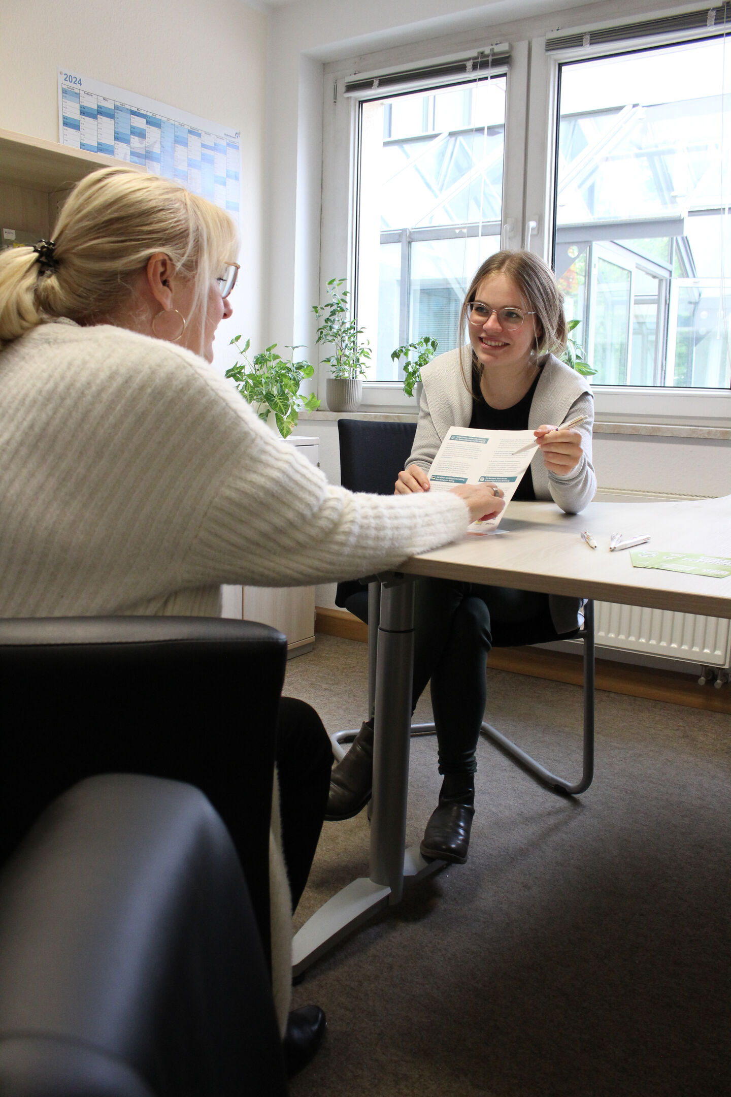A smiling woman is pointing at something on a sheet of paper, and a customer is sitting across from her